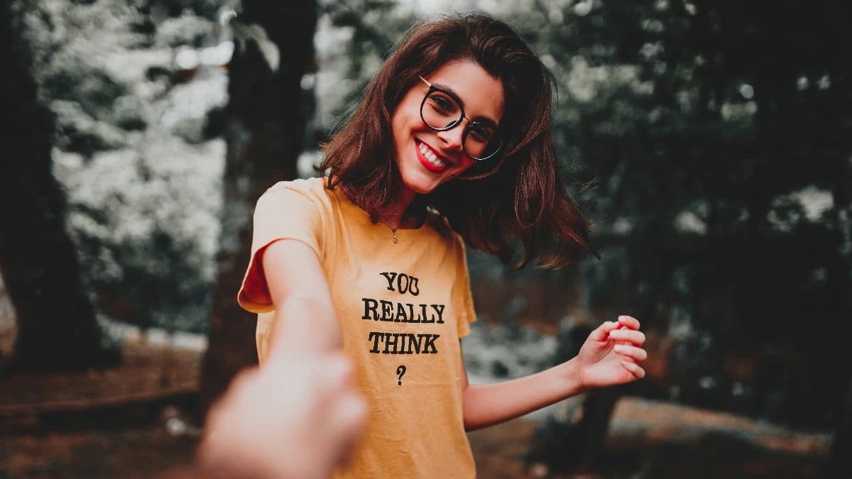 Cheerful woman in glasses holding hands outdoors, wearing a text t-shirt. via Pexels