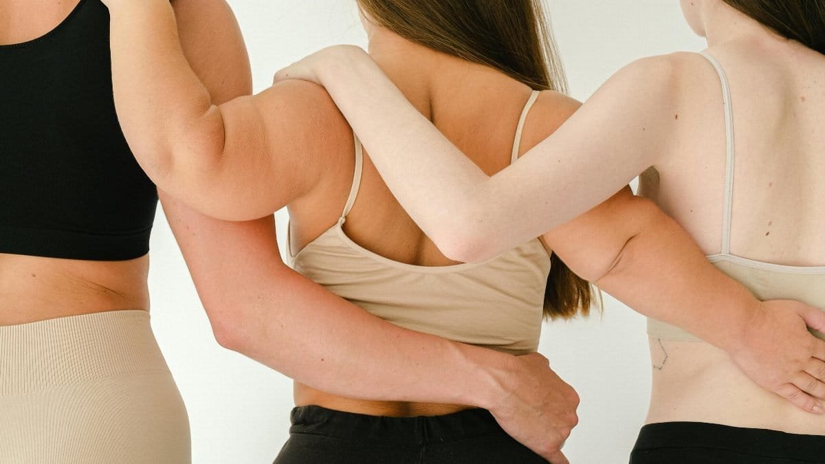 Three women standing together indoors, embracing body positivity and friendship. via Pexels