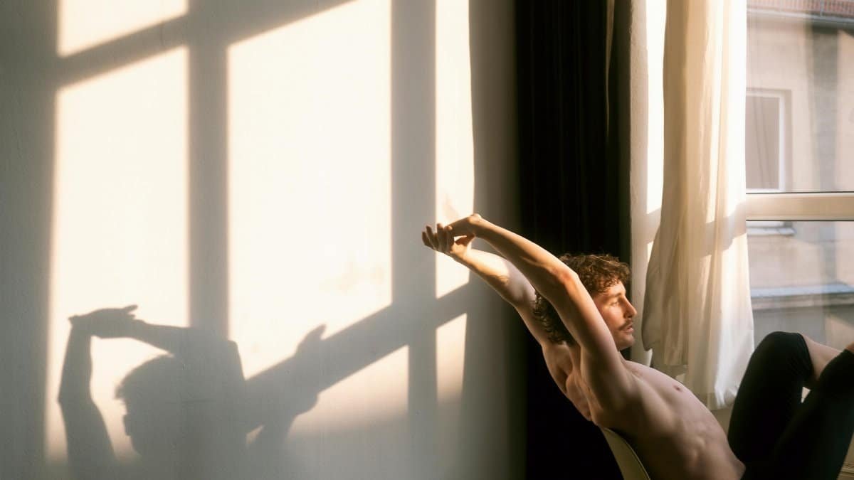 A man stretches in a sunlit room, casting a unique shadow on the wall. via Pexels