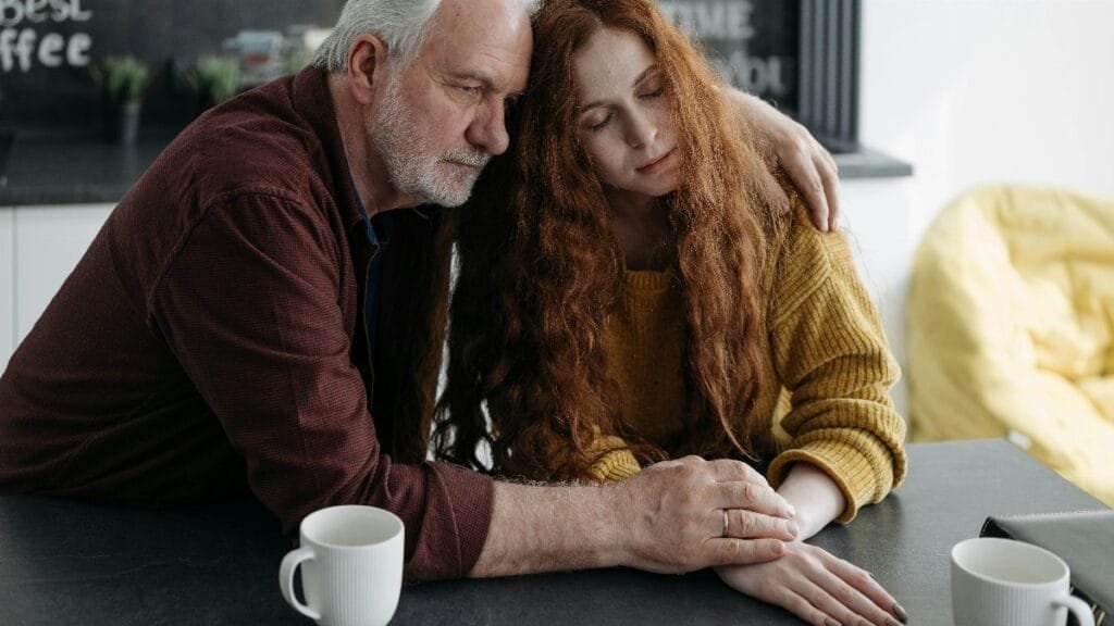 An elderly man consoles a young woman at a table in a warm, intimate indoor setting. via Pexels