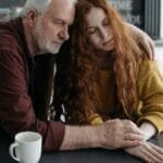 An elderly man consoles a young woman at a table in a warm, intimate indoor setting. via Pexels