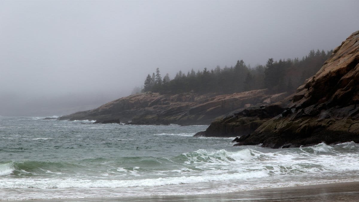 Foggy seascape featuring rocky cliffs, sandy beach, and gentle ocean waves. via Pexels