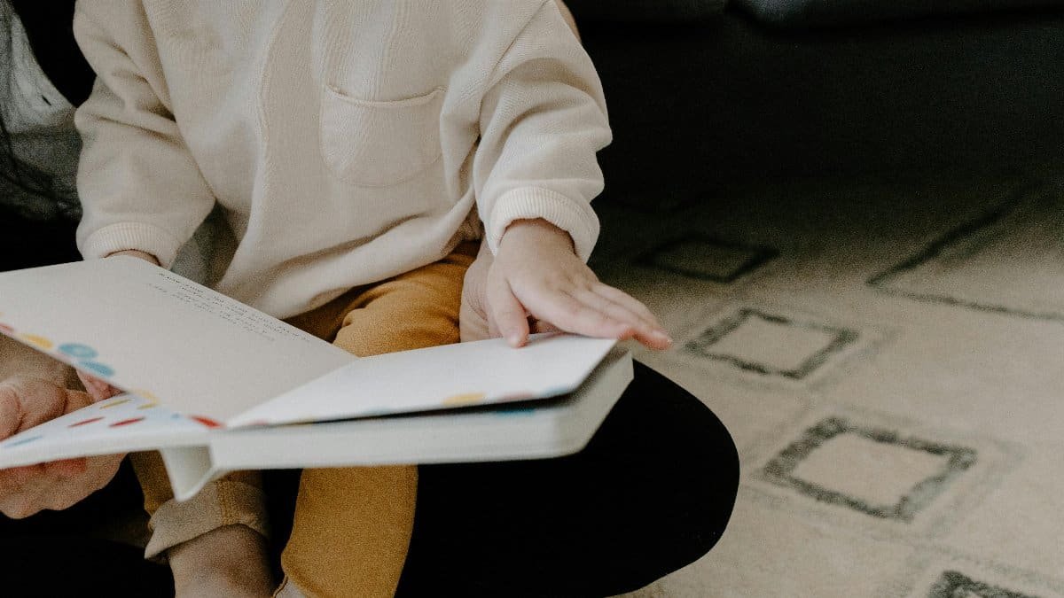 A young child sitting on a carpet, reading a colorful picture book indoors. via Pexels