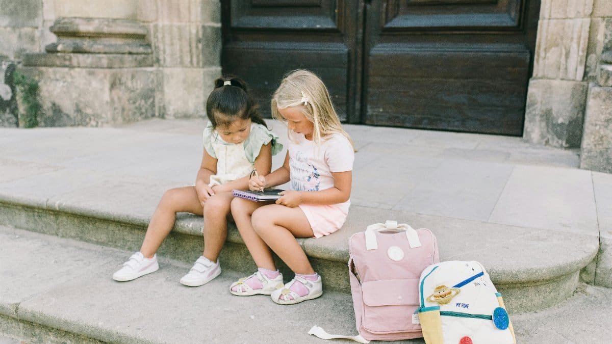 Two young girls sitting outside on steps, sharing a tablet with backpacks nearby. via Pexels