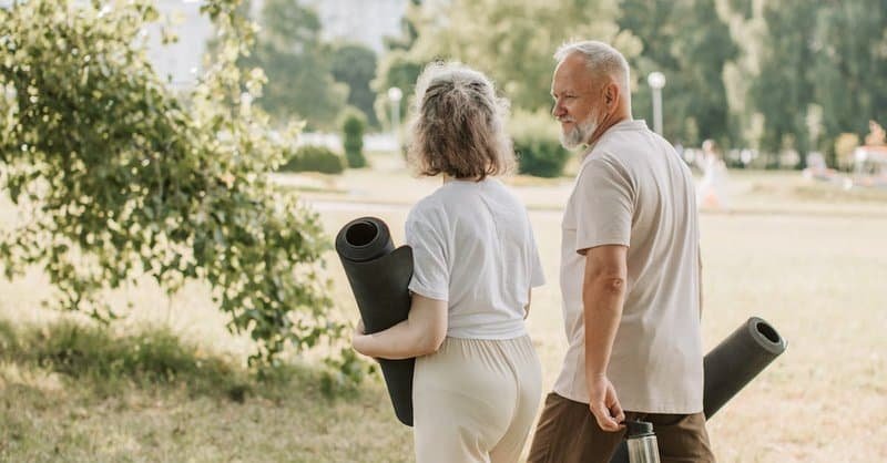 Elderly couple walking in park carrying yoga mats for a wellness session. via Pexels