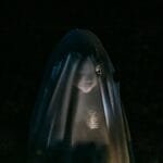 Mysterious little girl standing in darkness covered with white blanket as ghost and shining flashlight on face via Pexels