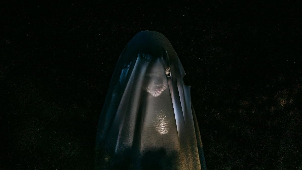 Mysterious little girl standing in darkness covered with white blanket as ghost and shining flashlight on face via Pexels