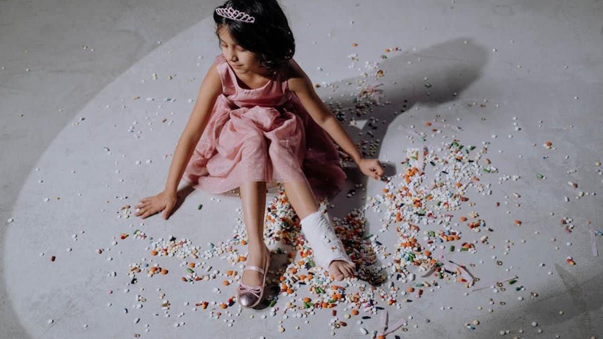 Asian girl in a pink dress with orthopedic cast, sitting on scattered pills. via Pexels