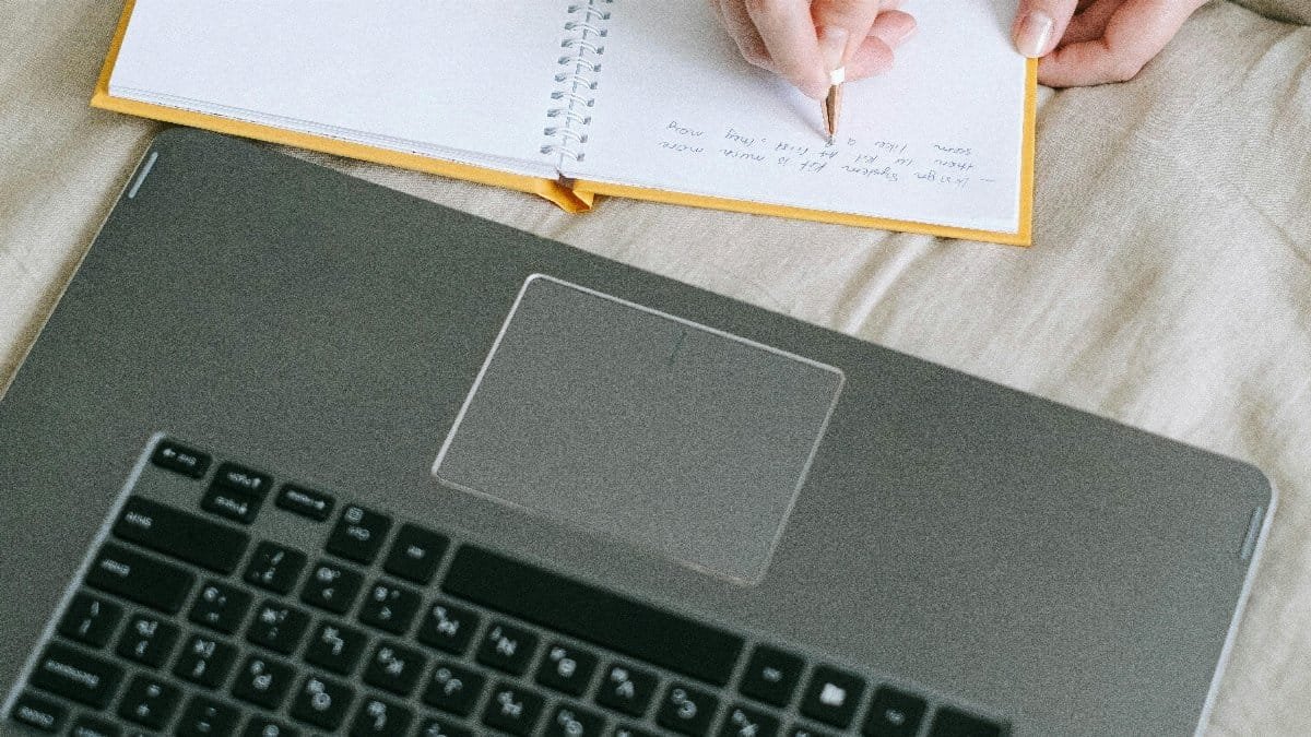 Close-up image of hands writing in a notebook beside a laptop, capturing a cozy learning environment. via Pexels