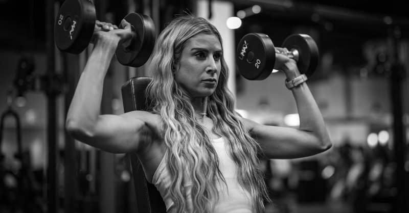 Focused woman working out with dumbbells on a weight bench in a gym setting. via Pexels