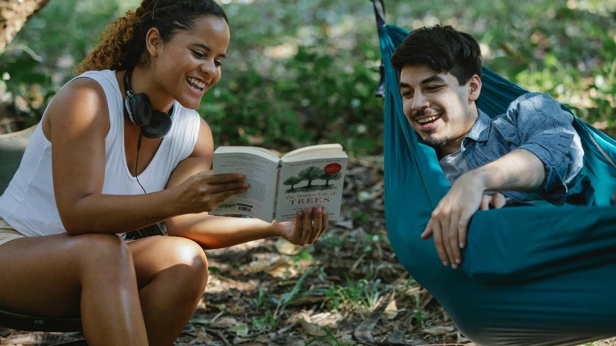 Happy couple laughing and reading a book together outdoors in a green hammock. via Pexels