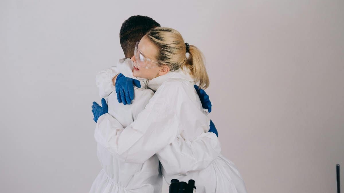 Two scientists hugging while wearing protective gear and goggles in a lab setting. via Pexels