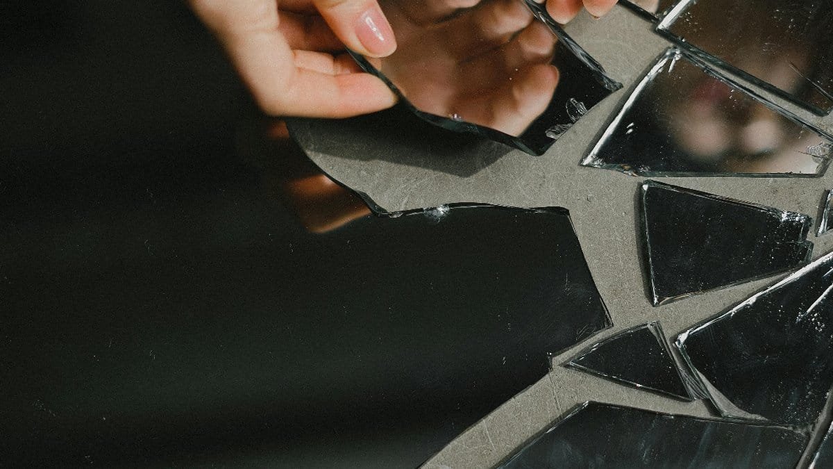 Close-up of hands touching reflective mirror shards on floor, symbolizing reflection and introspection. via Pexels