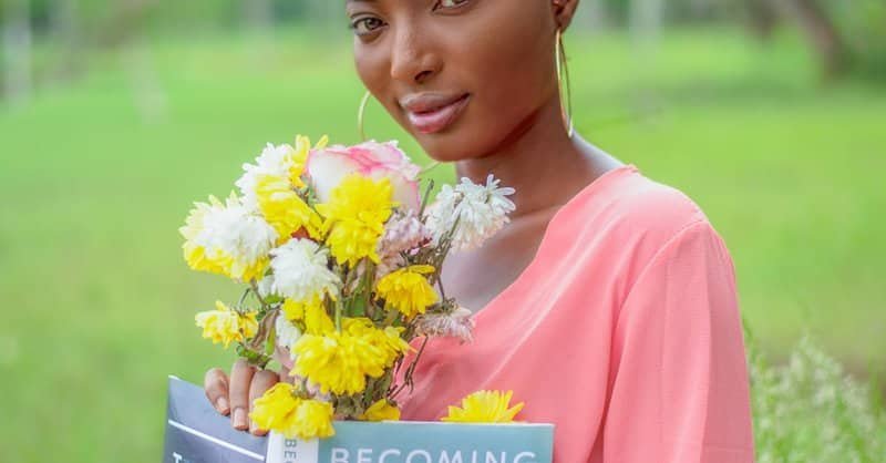 Smiling woman in pink dress holds bouquet and book 'Becoming' in a lush outdoor setting. via Pexels