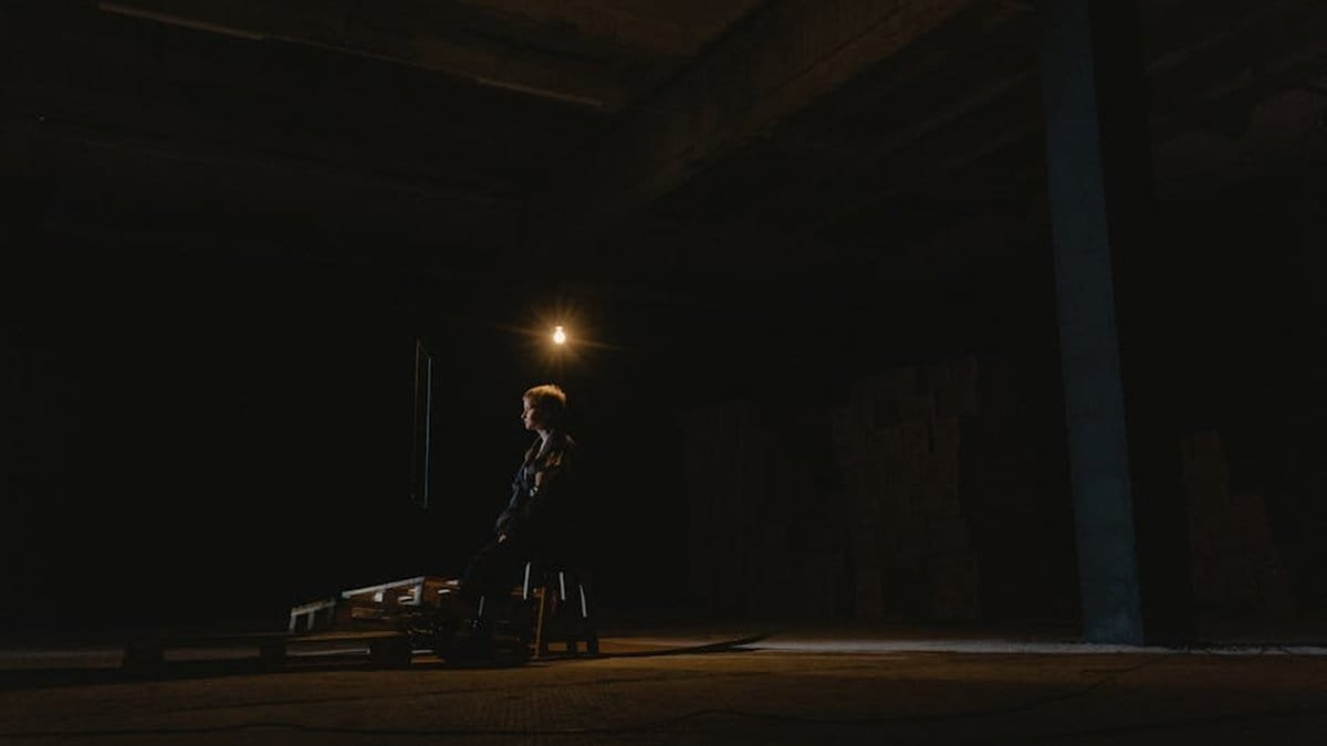 A solitary figure seated in a dark industrial space, illuminated by soft light. via Pexels