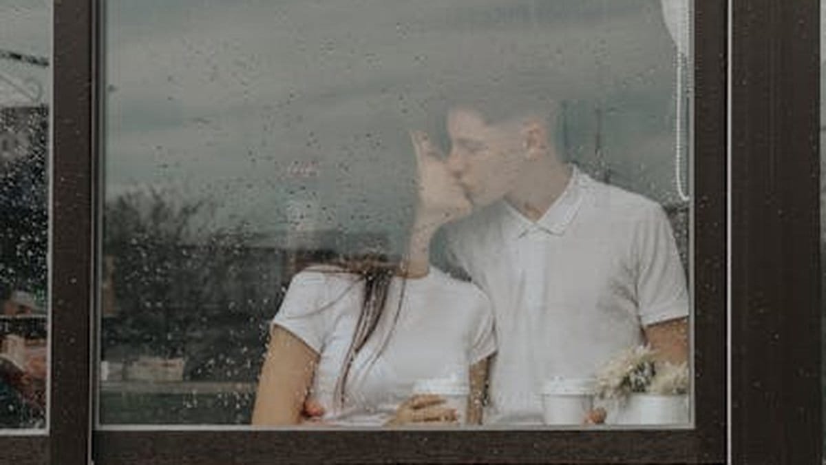 A young couple shares a tender kiss inside a café, viewed through a rain-spattered window. via Pexels