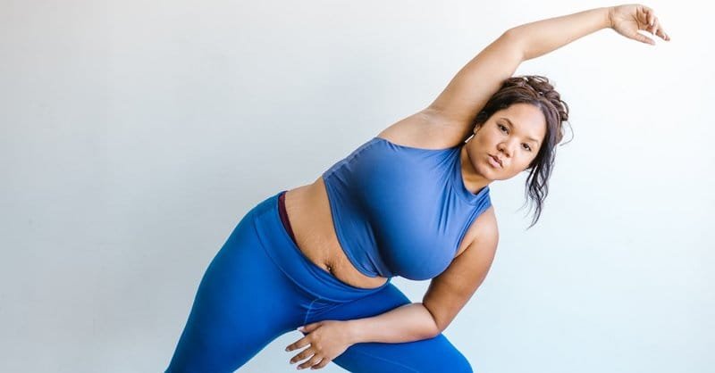 A confident plus size woman in blue activewear performing a stretching exercise indoors, promoting fitness and body positivity. via Pexels