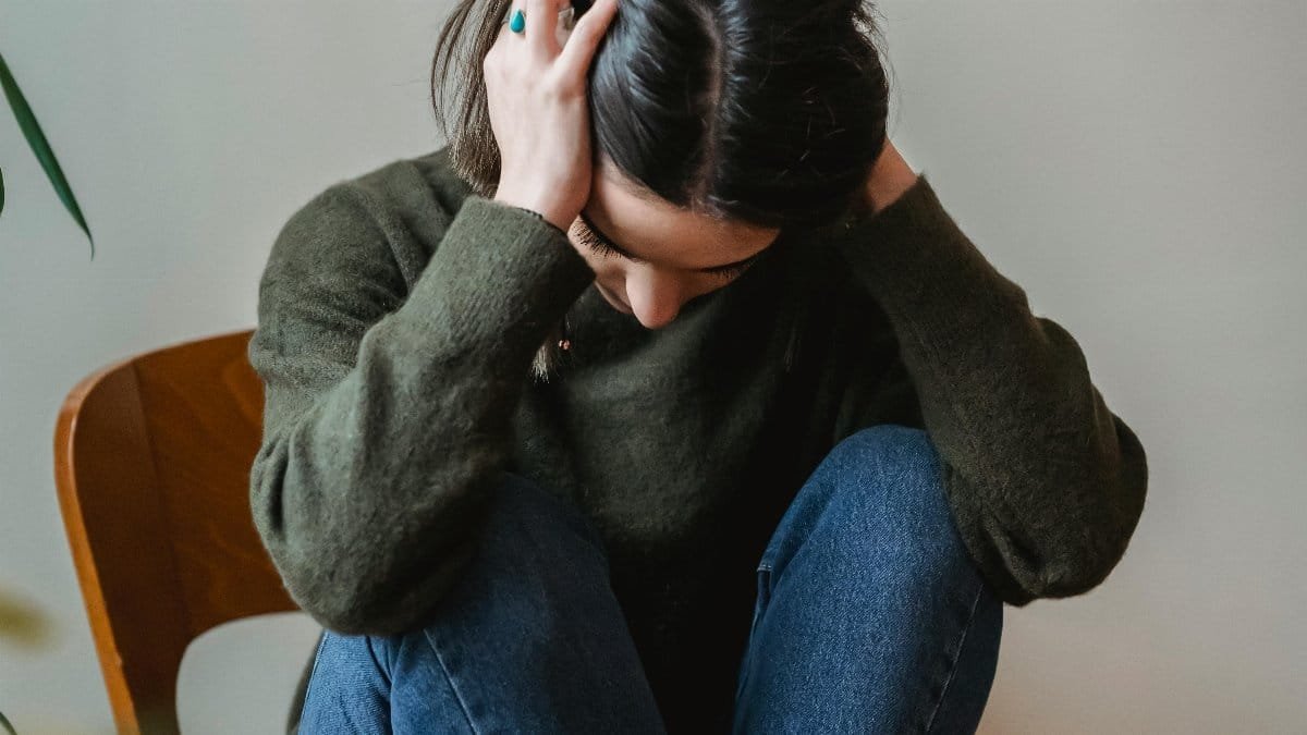 A young woman sitting on a chair indoors, holding her head in distress. via Pexels