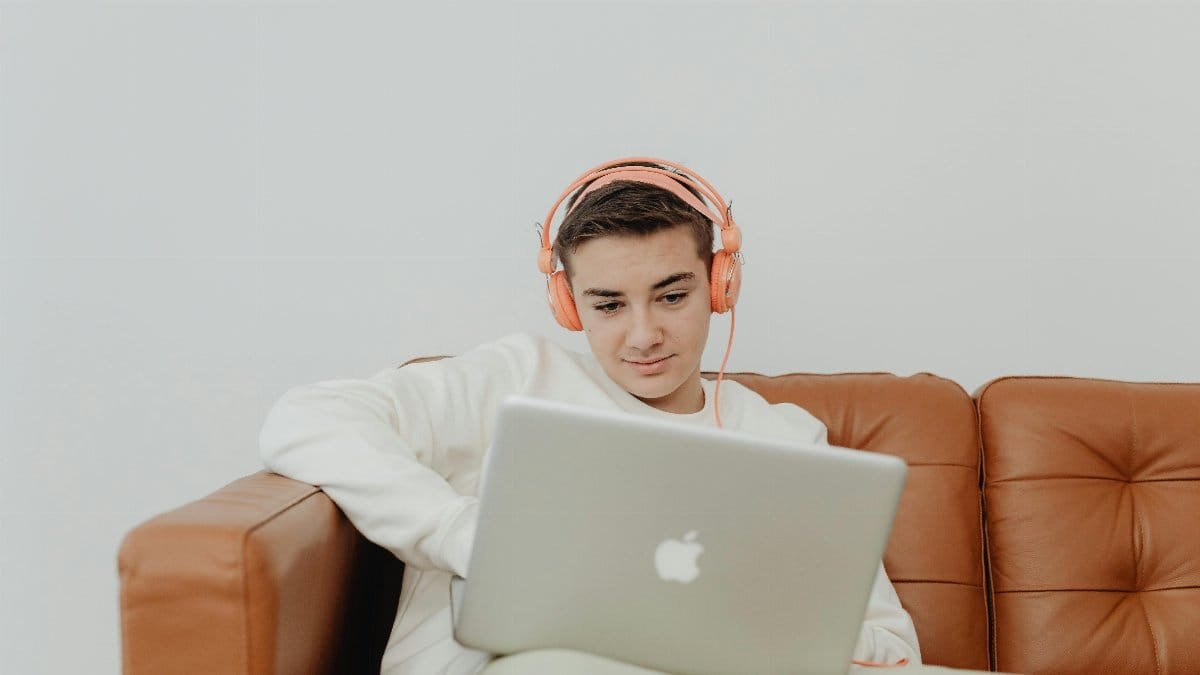 Young man sitting on a couch using a laptop with headphones, enjoying music indoors. via Pexels