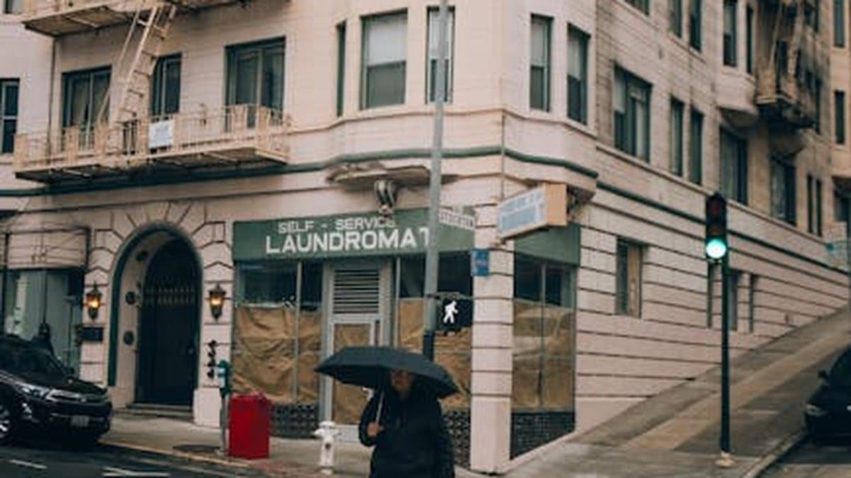 A man crosses a city street under an umbrella by a laundromat on a rainy day. via Pexels