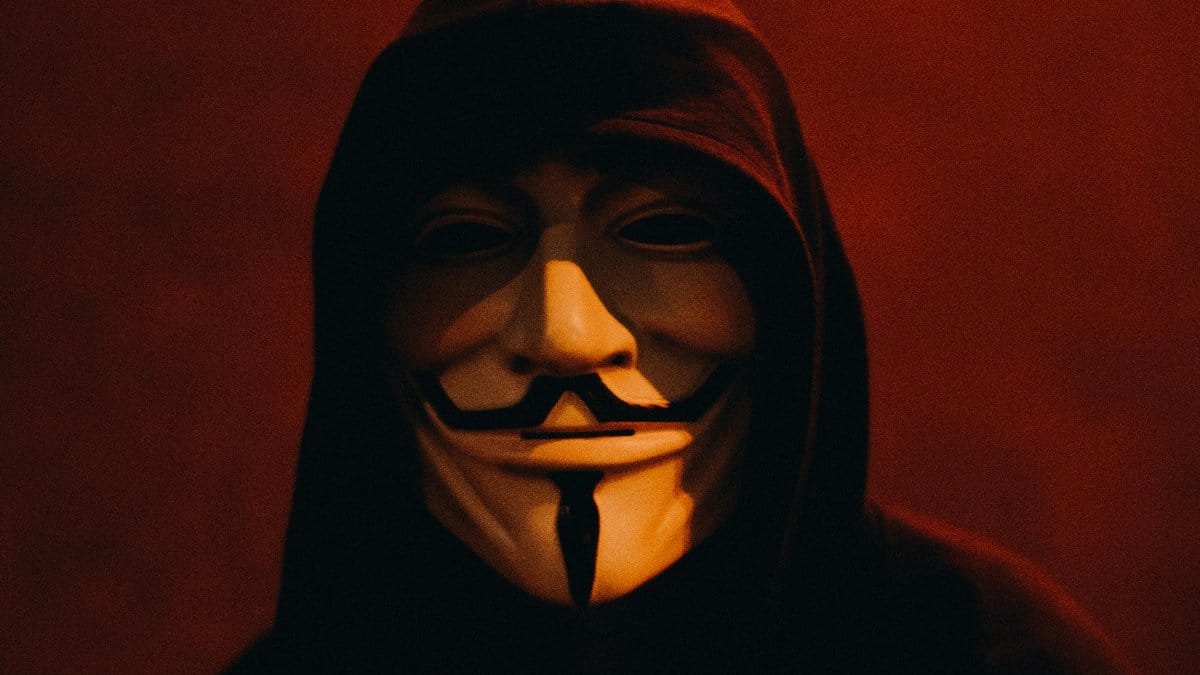 A person wearing a Guy Fawkes mask and hood in a dark, mysterious setting. via Pexels