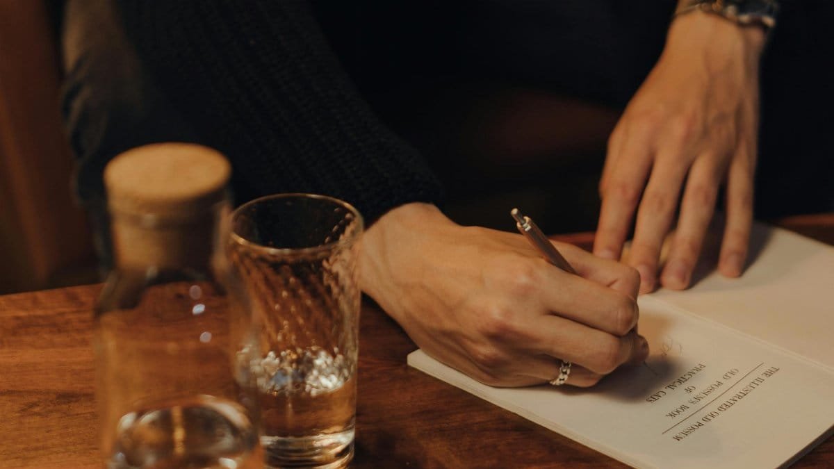 Person writing in a notebook on a wooden table with a glass of water. via Pexels