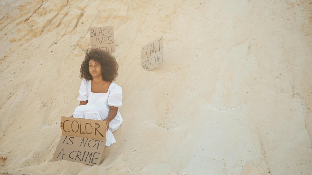 Woman in white dress holding sign 'Color is Not a Crime' on sand dunes, advocating for racial equality. via Pexels