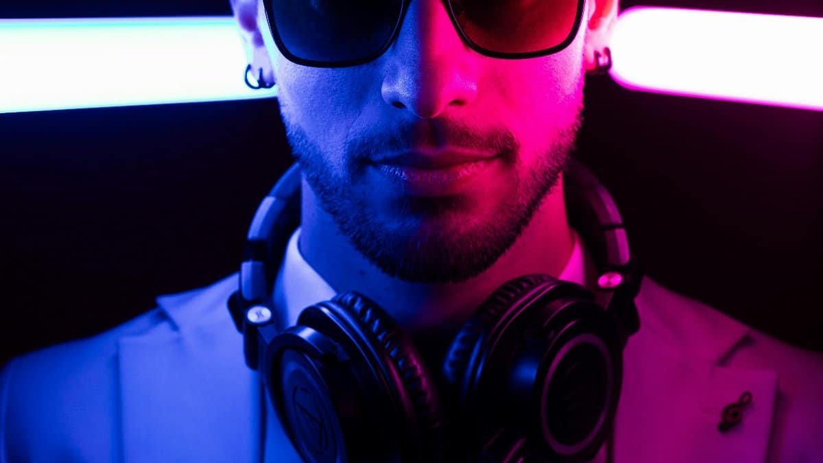 A stylish DJ wearing sunglasses and headphones illuminated by neon pink and blue lights. via Pexels