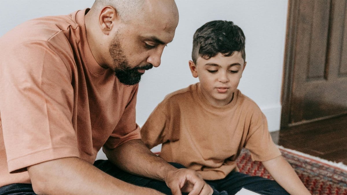 A father and son bonding while reading a religious text on a rug indoors. via Pexels
