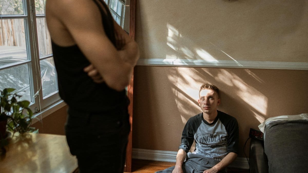 Two young men in a tense indoor setting, one standing with arms folded, the other sitting on the floor. via Pexels
