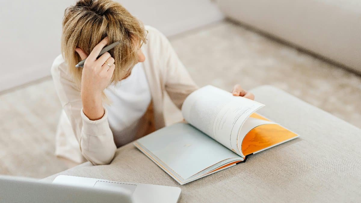 Blonde woman studying in living room with a book and laptop, creating a relaxed atmosphere. via Pexels