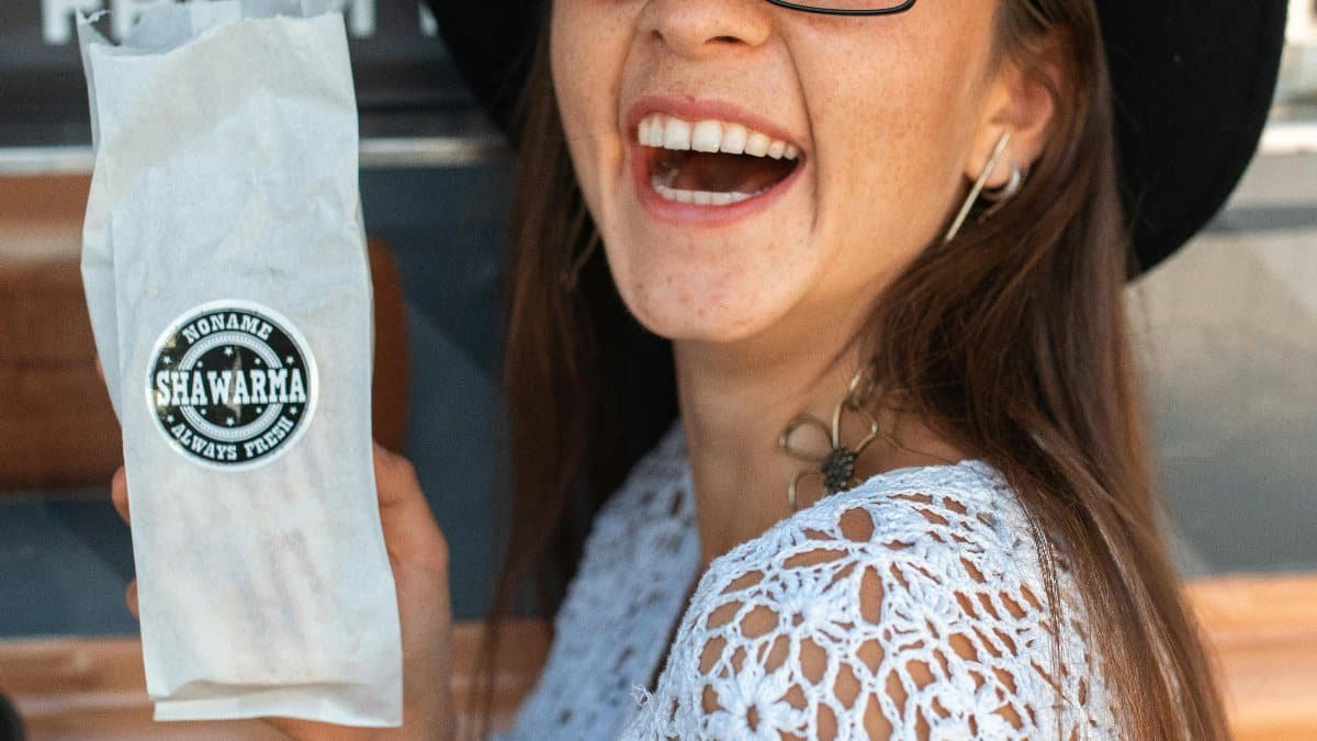 Cheerful woman showcasing shawarma while smiling, wearing a white top and black hat. via Pexels