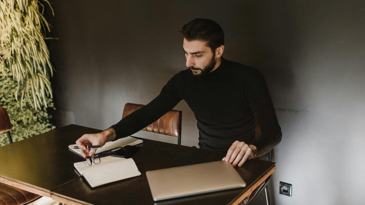 Adult man sitting at a desk working with a laptop, notebook, and pen indoors. via Pexels