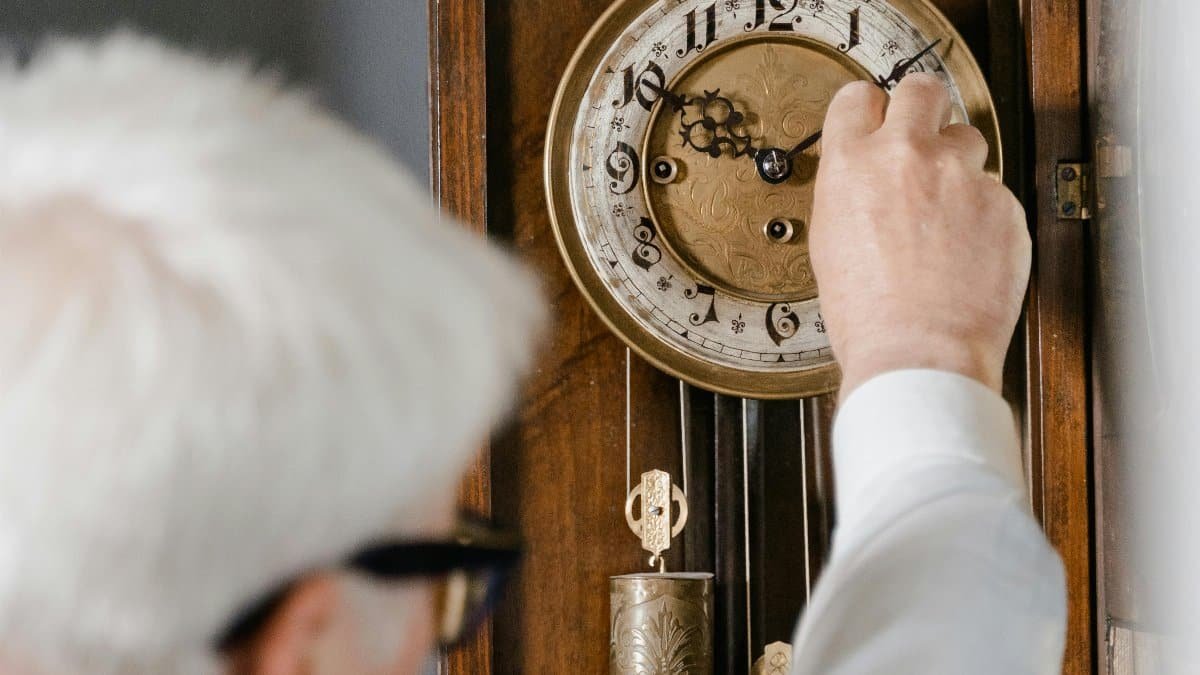 An elderly man adjusting a vintage wooden clock in an indoor setting. via Pexels