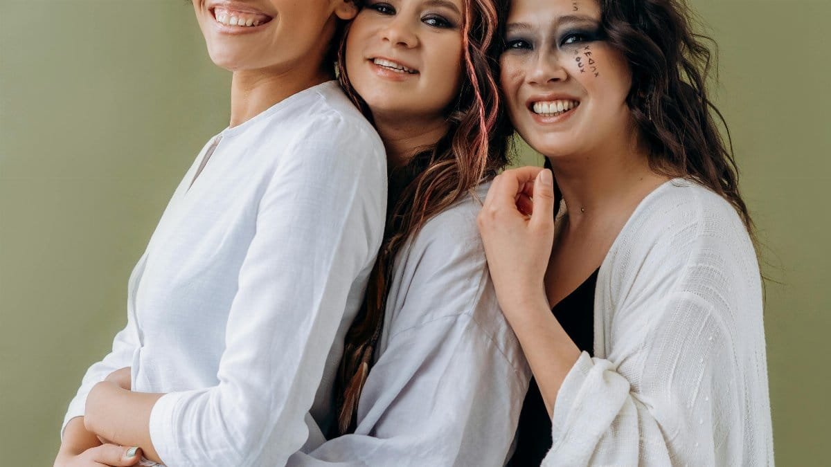 Three multicultural women in white attire stand happily embracing against a green backdrop. via Pexels