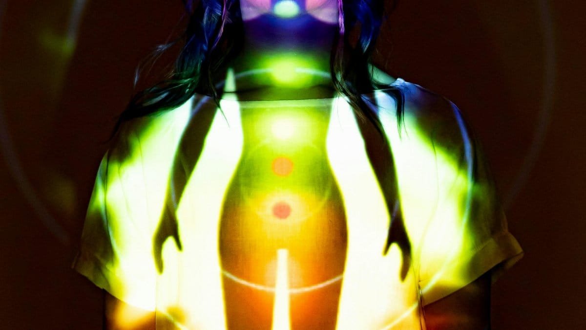 A woman stands in a dark room with a colorful chakra projection on her body. via Pexels
