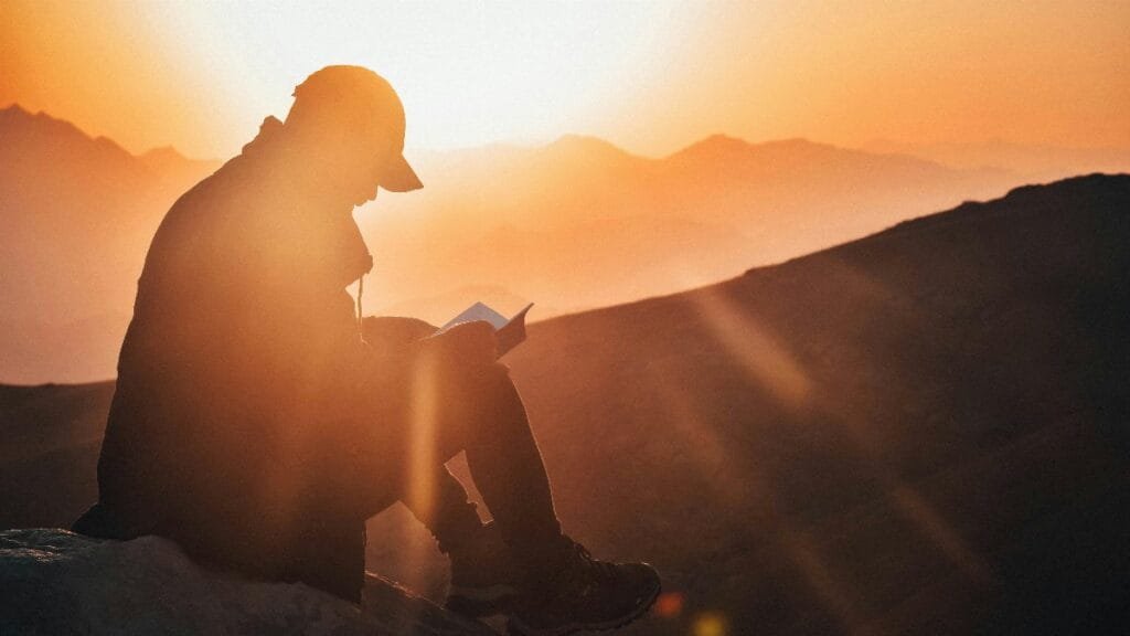 A man reads a book silhouetted against a vibrant sunset, seated on a mountain edge. via Pexels