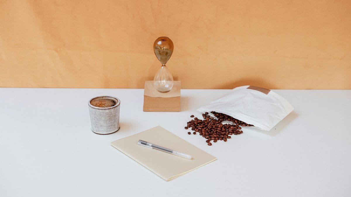 A minimalist workspace featuring coffee beans, an hourglass, a notebook, and a pen for creative inspiration. via Pexels