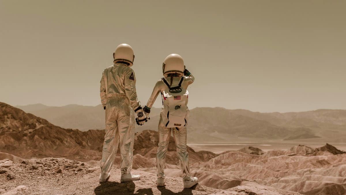 Two astronauts in space suits holding hands in a desert, Mars-like terrain. via Pexels