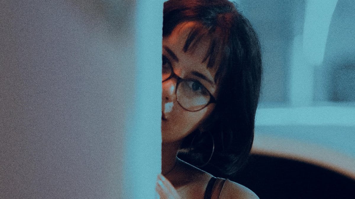 A woman with glasses and short hair peeks from behind a wall in a mysterious indoor setting. via Pexels