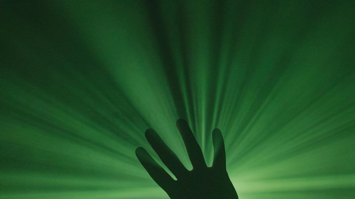 A mysterious silhouette of a hand reaching toward vibrant green light rays creating a dynamic visual effect. via Pexels
