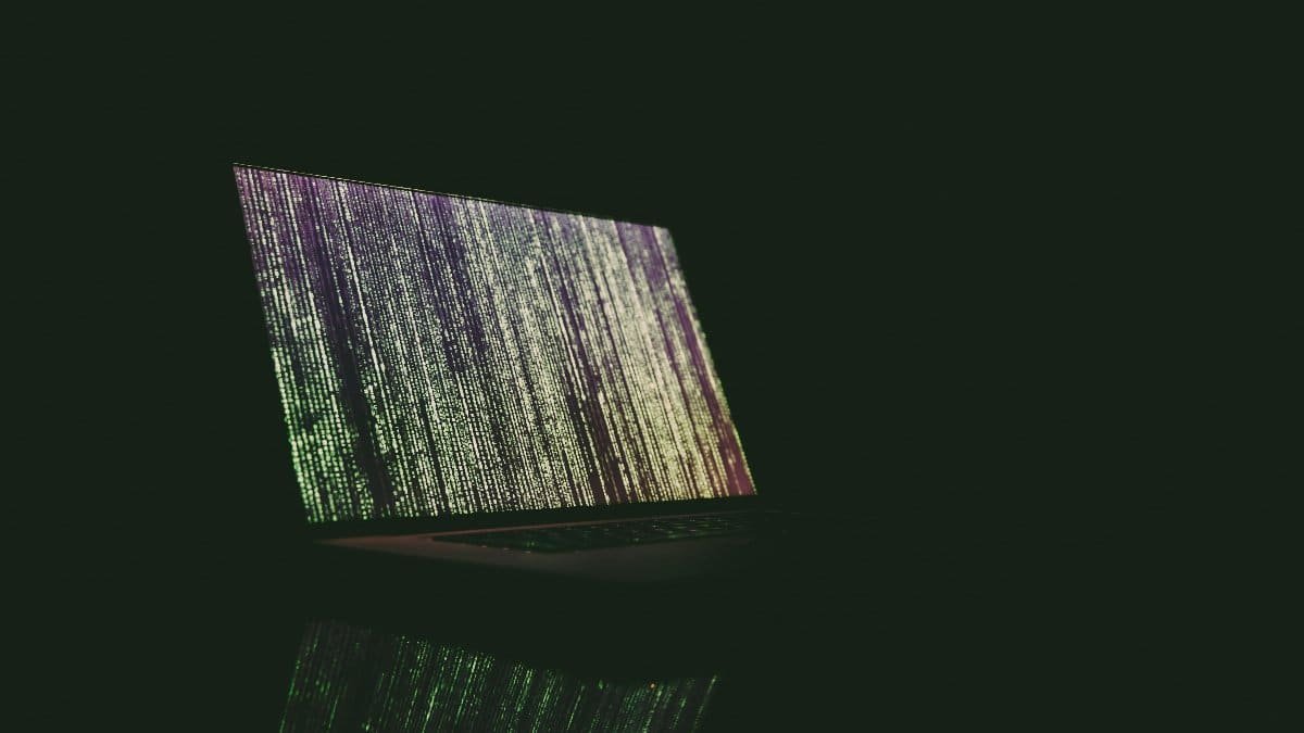 A laptop displaying green and purple code in a dark room, reflecting on a surface. via Pexels