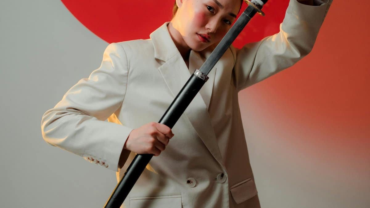 Striking portrait of an elegant woman in a blazer, confidently posing with a katana sword against a bold red circle background. via Pexels