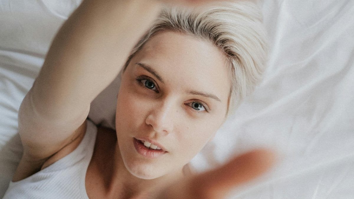 Portrait of a young woman in a white tank top lying on a bed with white linen, reaching towards the camera. via Pexels