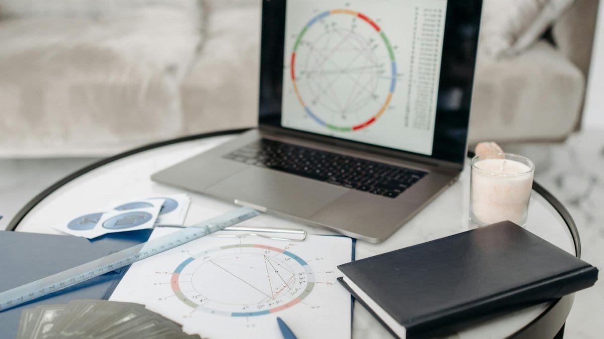 Astrology charts displayed on a laptop next to tarot cards and notebooks on a table.