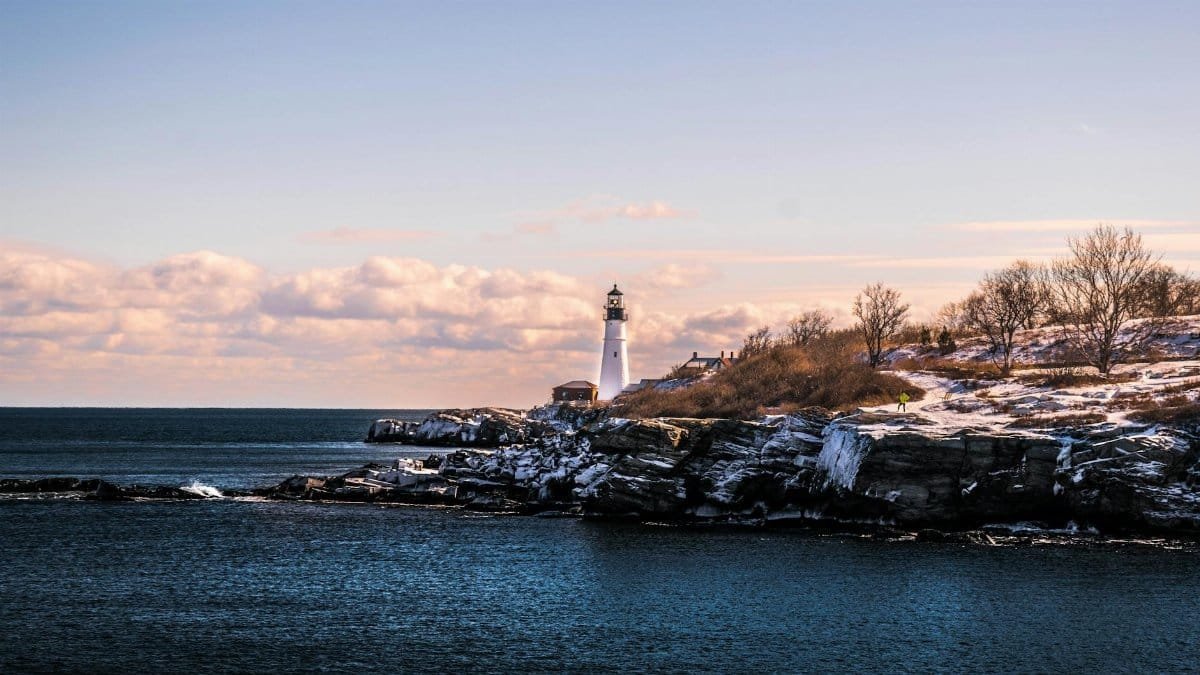 A picturesque winter view of a lighthouse on the rocky Maine coastline under a bright sky.