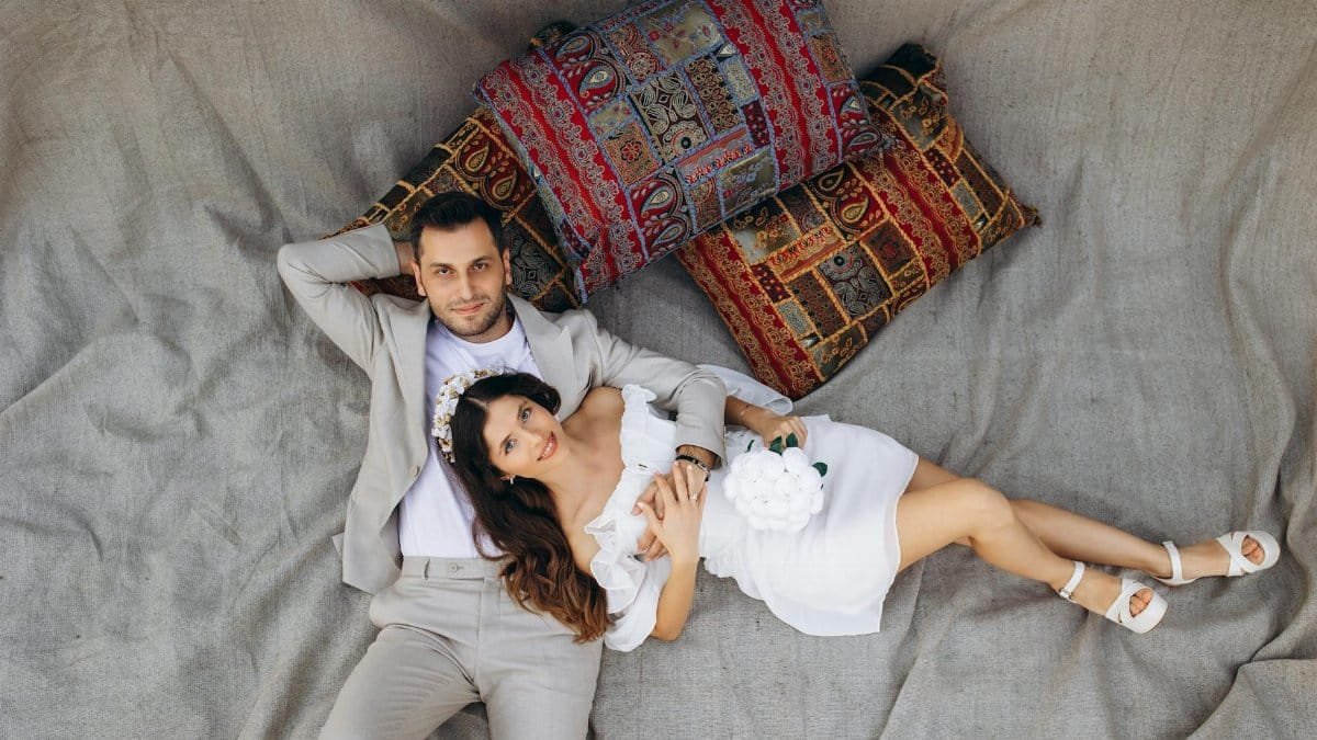 A couple wearing formal attire relaxing on a carpet with colorful pillows, captured from above.