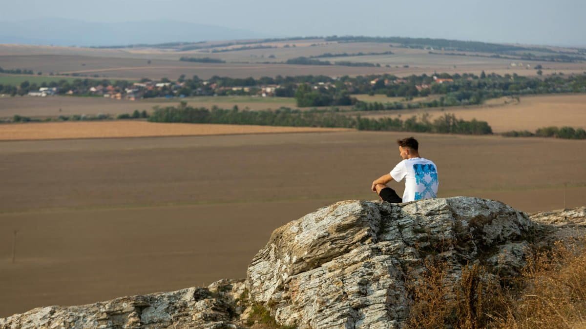 A man sits on a rock overlooking the vast countryside in a serene outdoor setting.