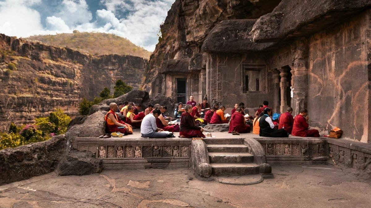 Group of monks in traditional robes meditating at an ancient rock temple during the day.
