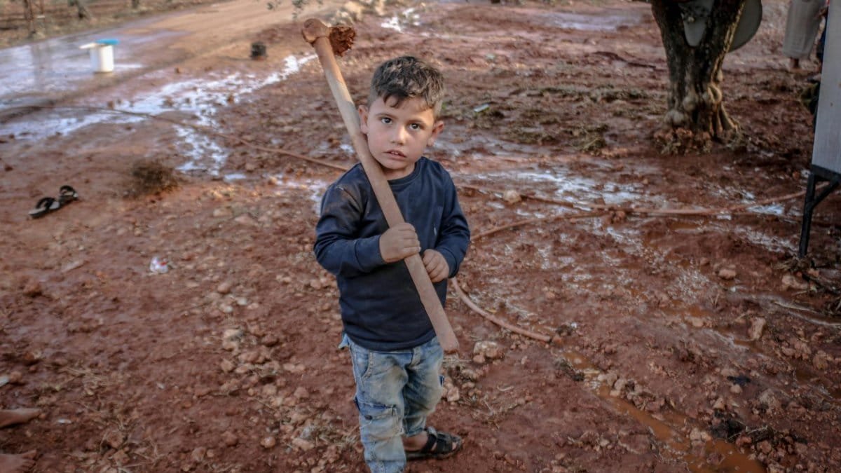 A young boy holding a stick stands on muddy ground in rural Idlib, Syria.